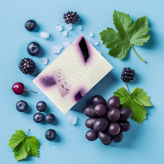 Savon au lait de brebis et trois beurres à la fragrance de raisin et bleuet entouré de raisins noirs, bleuets, mûres et feuilles vertes sur un fond bleu clair.